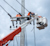 Three lineworkers in orange perform maintenance on a utility pole. One is in a bucket truck with an orange boom. Overcast sky.