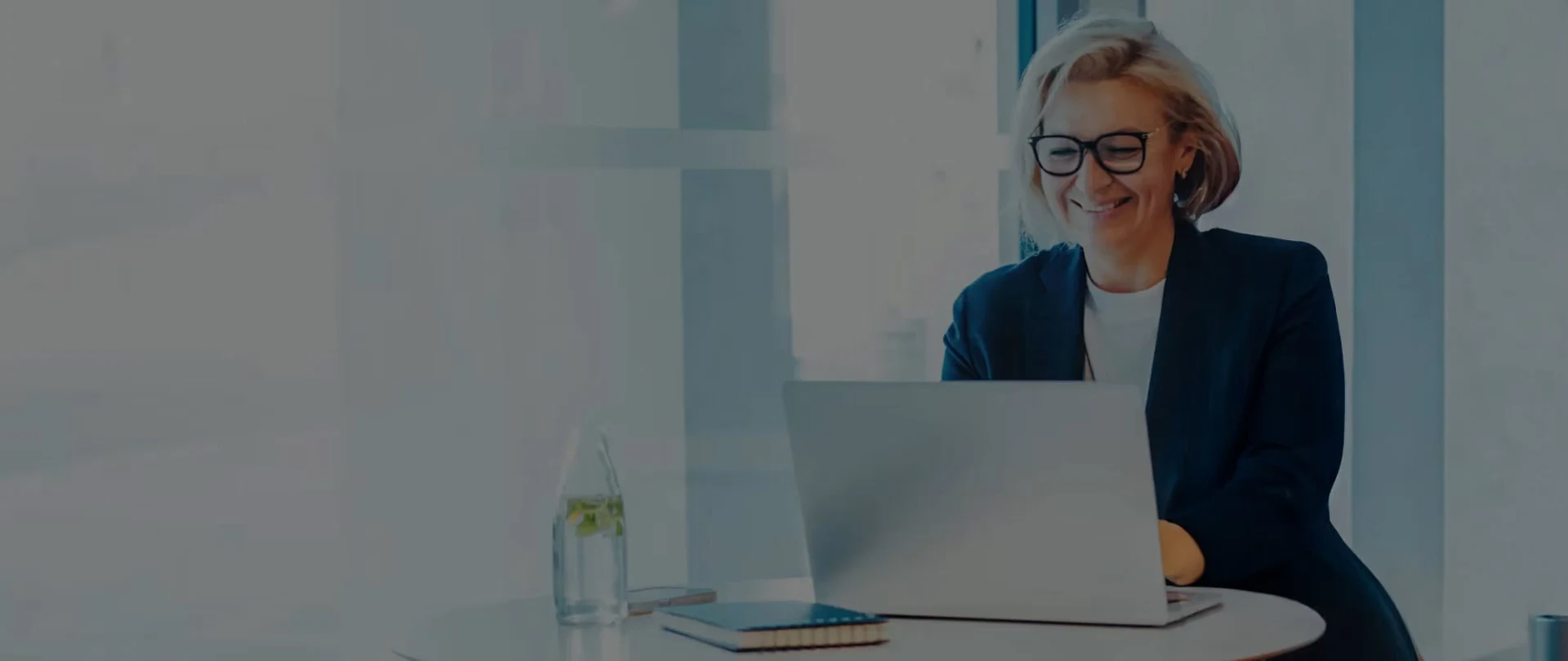 A smiling woman with short blonde hair and glasses sits at a table, looking at a laptop.