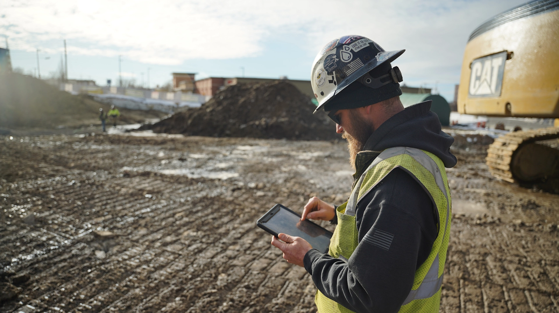 A construction worker uses a rugged Trimble tablet for site management at a muddy construction site near a CAT excavator.