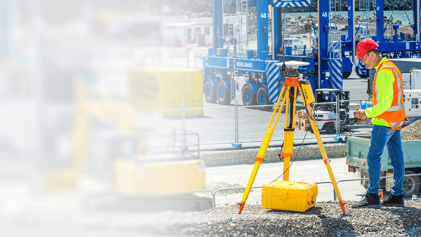 Surveyor on a construction jobsite with machinery behind them, standing next to the Trimble R750 Model 2 modular GNSS receiver mounted on a tripod with an antenna.