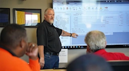 An instructor points to a construction estimating software interface on a large wall-mounted screen during a training session with several attendees.