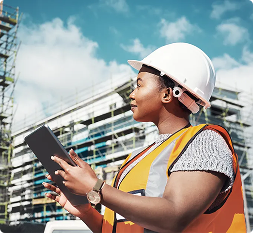 A person wearing a white hard hat and an orange high-visibility vest is holding a tablet while standing in front of a construction site.