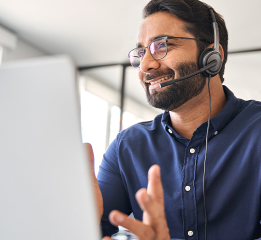 A smiling man wearing a headset and glasses while looking at a laptop, providing customer support.