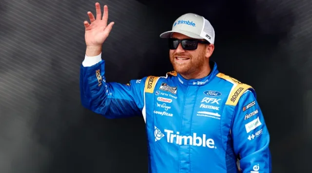 Chris Buescher, RFK Racing car driver in a blue and white racing suit and hat featuring Trimble branding waves against a dark background.