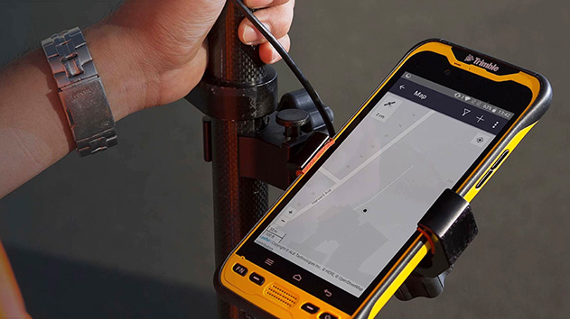 A person's hand is visible holding onto a pole, with a ruggedized, yellow and black smartphone or handheld device mounted to it.