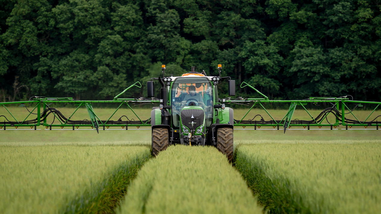 A head-on shot of a large, green agricultural tractor with wide sprayer booms extended, driving down a track in a field of tall, green crops.