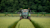 A head-on shot of a large, green agricultural tractor with wide sprayer booms extended, driving down a track in a field of tall, green crops.