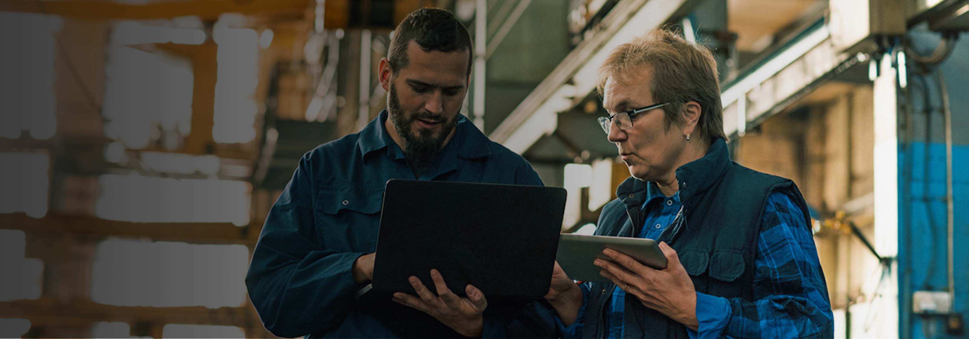 Two warehouse workers reviewing data on a laptop and a tablet.
