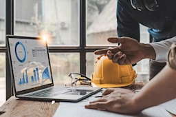 A person points at construction data charts on a laptop next to a yellow hard hat and safety glasses in an office setting.