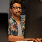 A man in a plaid shirt and glasses sits at a desk, smiling while looking at a computer monitor.