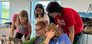 Group of children & young adults huddled around a boy working on laptop in classroom