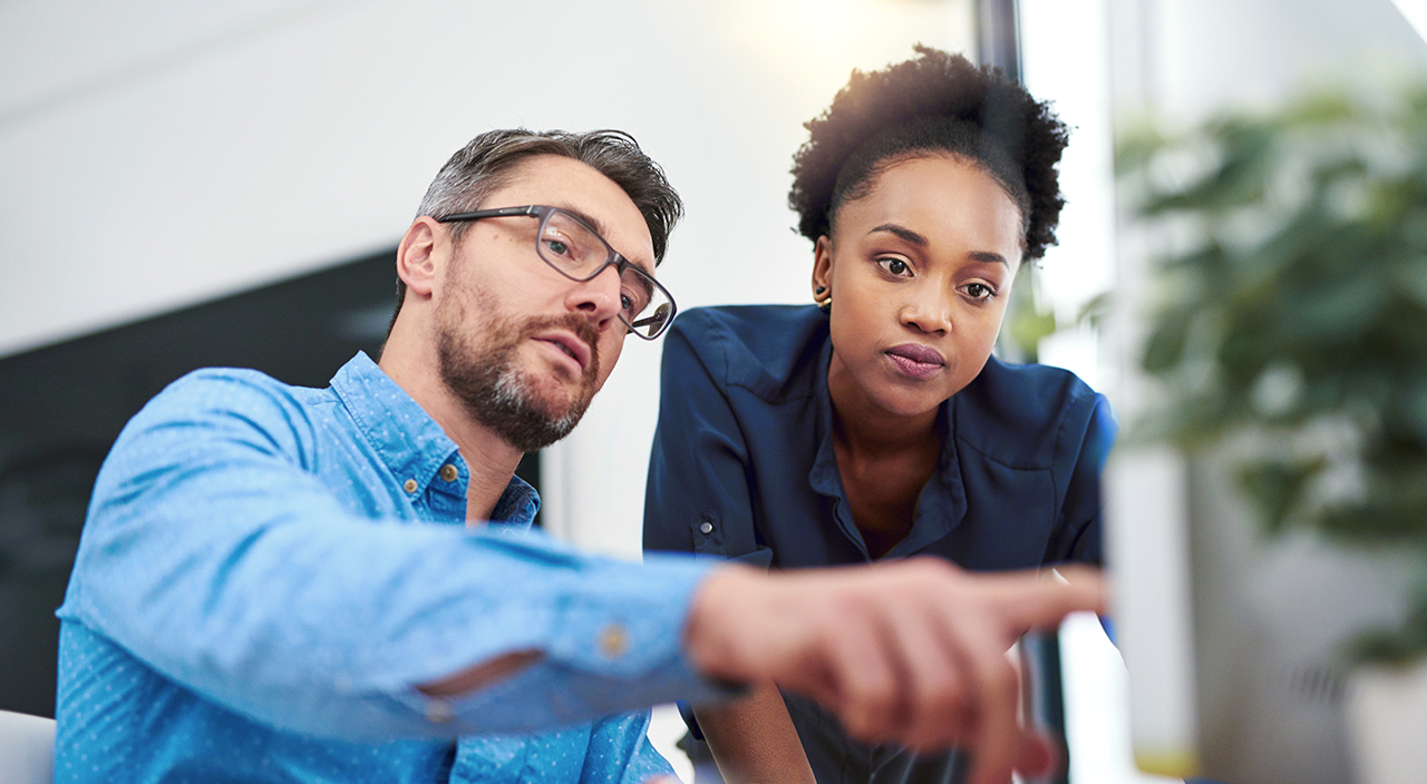 A man and a woman in an office look at a computer screen together as the man points to a detail during a collaborative discussion.