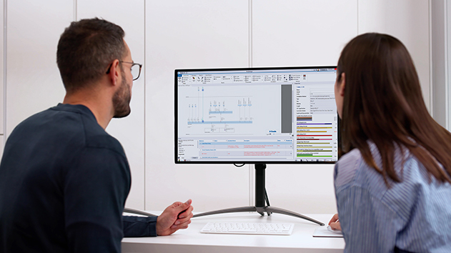 Two people view a computer monitor displaying Trimble Electrical Designer software with circuit diagrams and electrical compliance data charts.