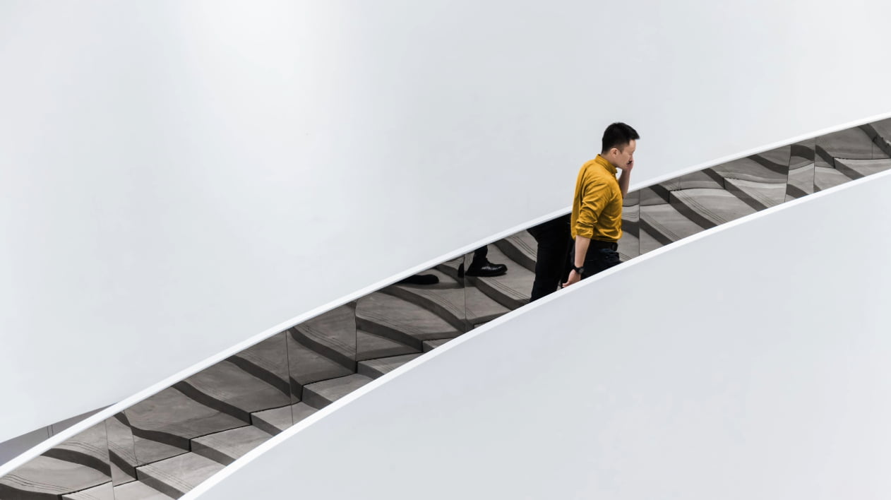  A man in a yellow shirt walks up a minimalist, reflective escalator beside a plain white wall.
