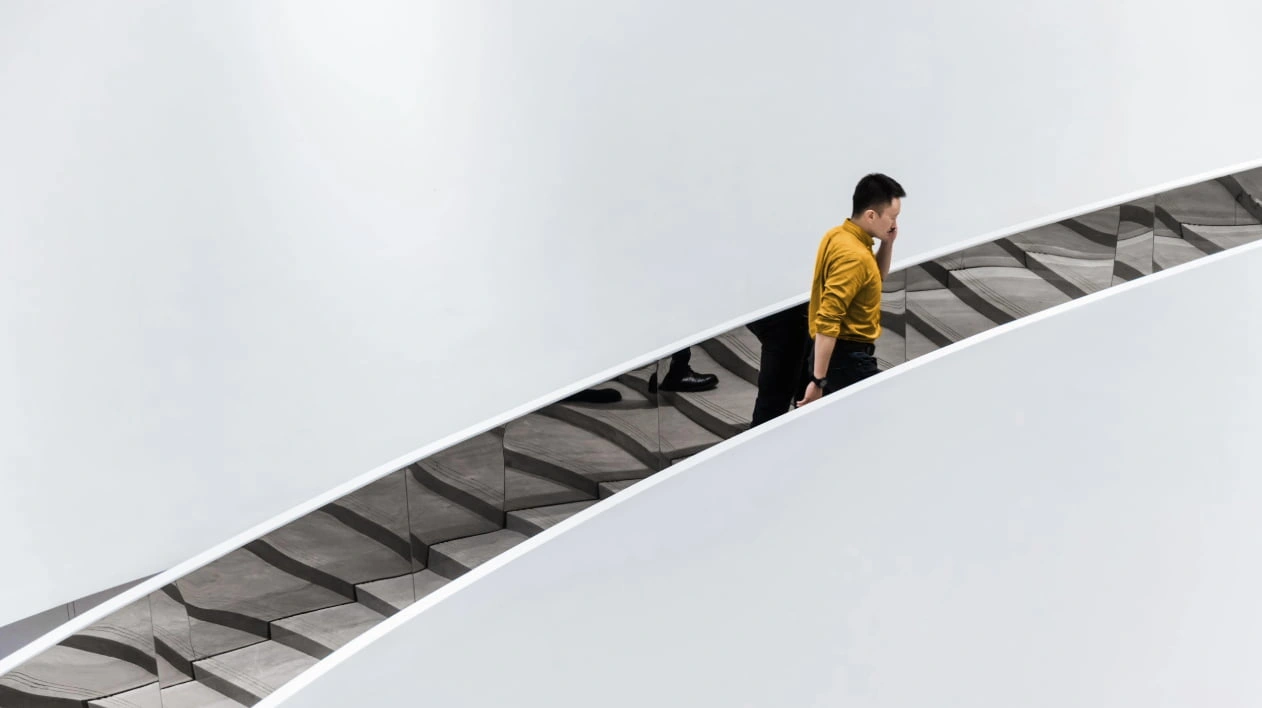 A man in a yellow shirt walks up a minimalist, reflective escalator beside a plain white wall.