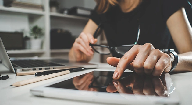 A woman at a desk touches a tablet screen while holding eyeglasses, with a laptop and writing utensils nearby.