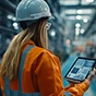 A worker wearing a white hard hat and an orange high-visibility jacket uses a tablet in a blurred industrial factory setting.