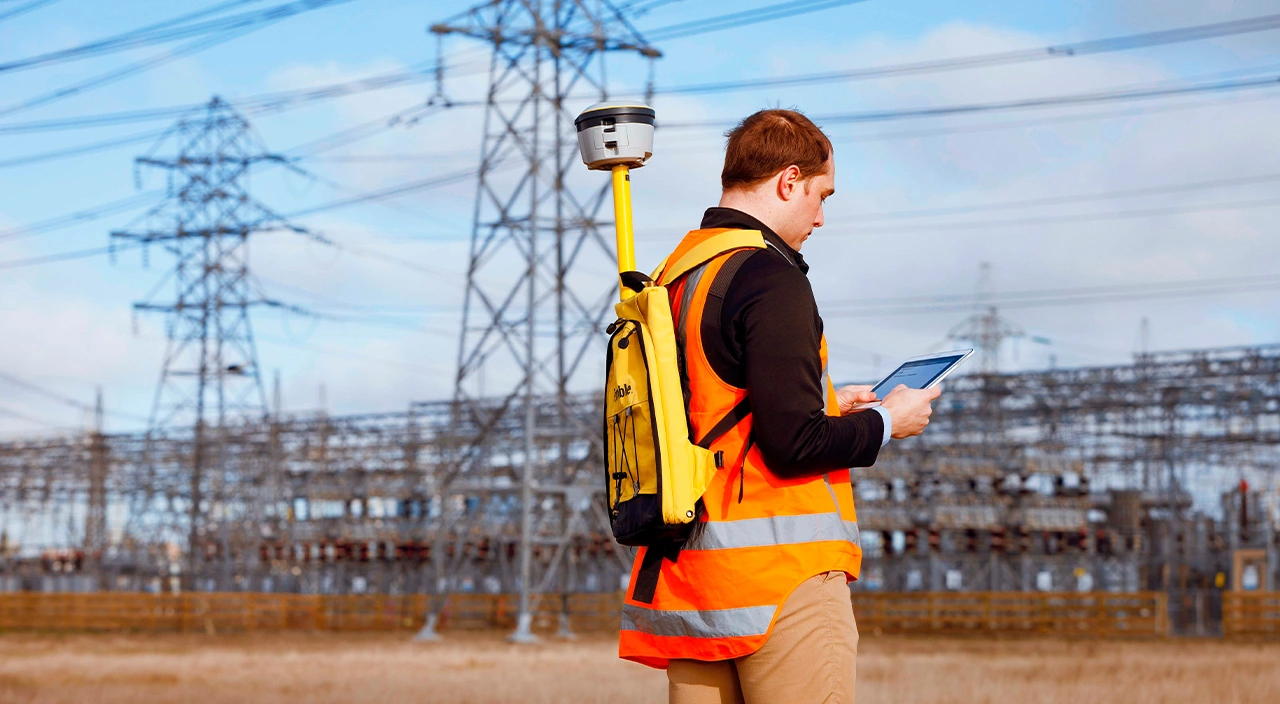 A worker in an orange vest with a Trimble geospatial device looks at a tablet near power lines.