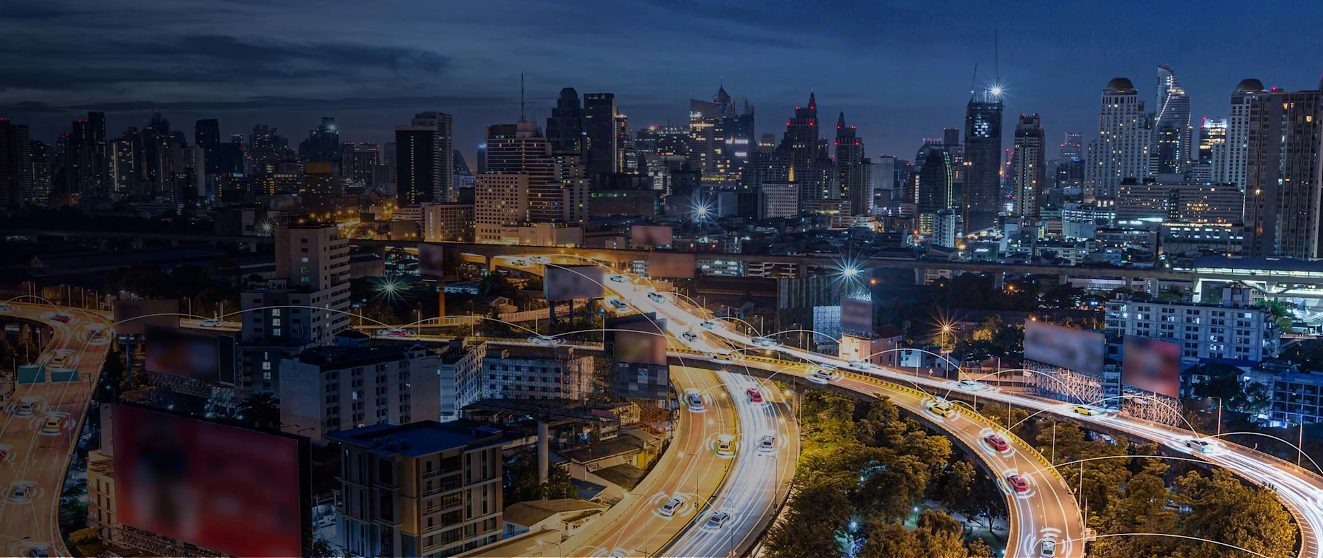 An aerial, nighttime long exposure shot captures a bustling highway interchange in a modern city, with the brightly lit roadways winding through a dense urban landscape of tall buildings.