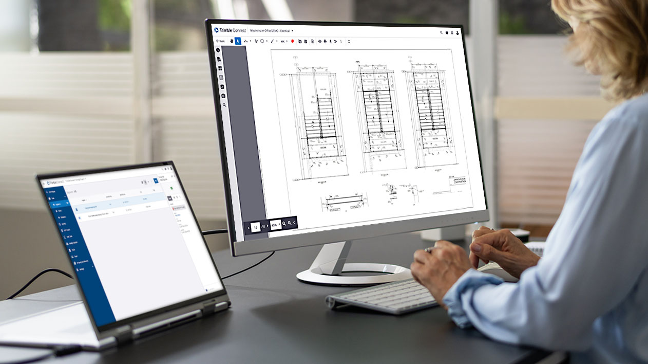 A professional uses Trimble Connect on a laptop and desktop monitor to review digital construction drawings and electrical plans.