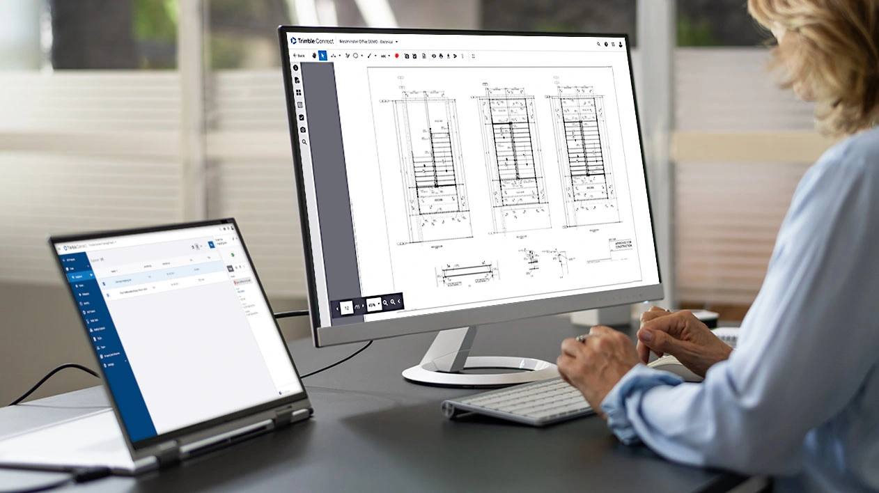 A professional uses Trimble Connect on a laptop and desktop monitor to review digital construction drawings and electrical plans.
