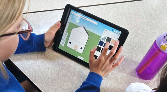Jeune étudiant assis à un bureau utilisant une tablette numérique pour créer un modèle de maison en 3D à l’aide d’un logiciel de conception.