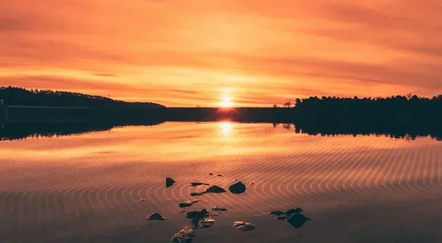 Golden sunset over massive dam, reflecting brightly on vast reservoir with concentric ripples. Dam silhouetted against sky.