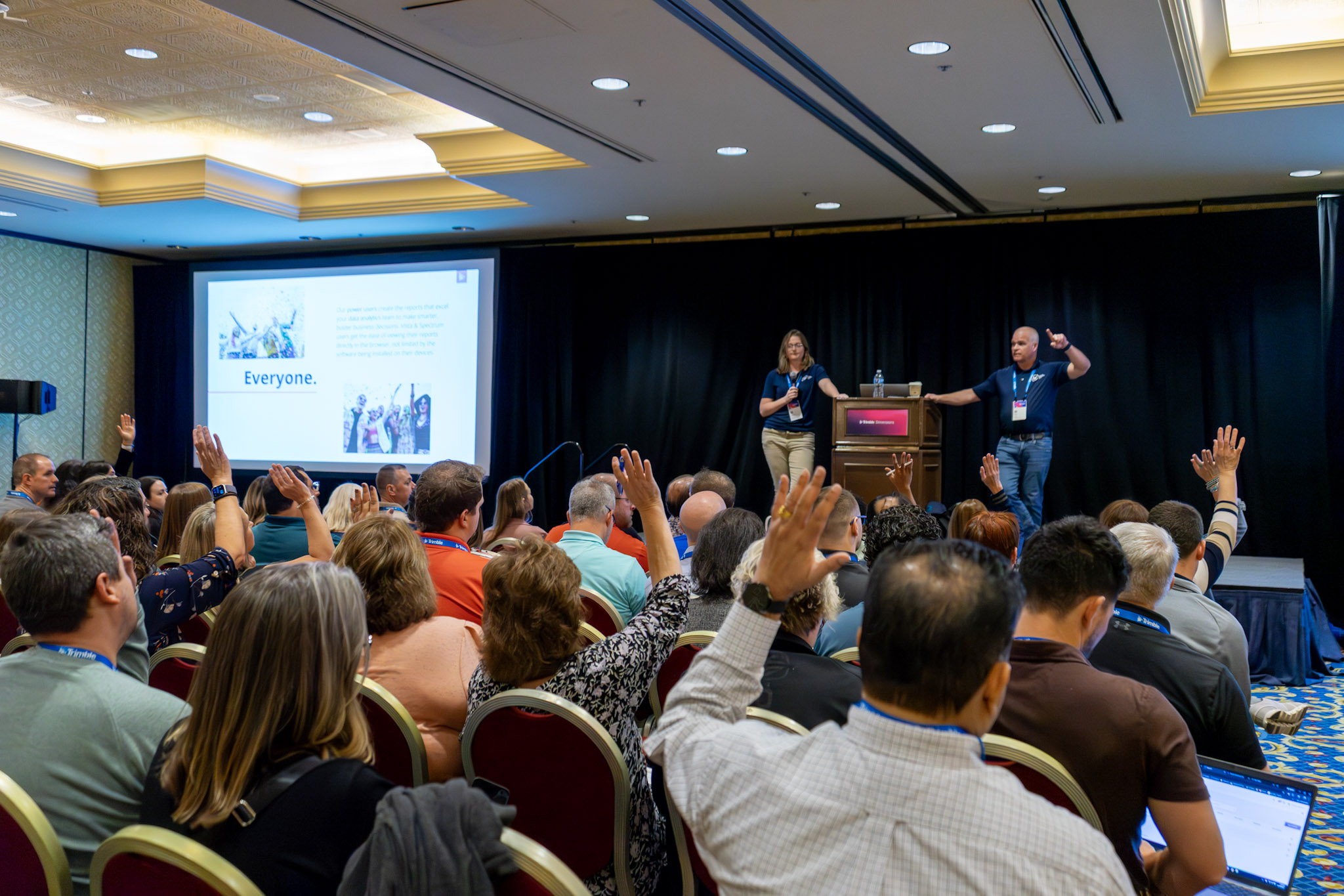 An audience raises their hands during a presentation in a conference room. Two speakers stand on a stage with a podium in front of a black backdrop, next to a large projection screen displaying a slide with text and photos.