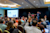 An audience raises their hands during a presentation in a conference room. Two speakers stand on a stage with a podium in front of a black backdrop, next to a large projection screen displaying a slide with text and photos.