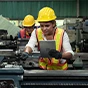 A factory worker in a yellow hard hat and safety vest uses a Trimble tablet in an industrial workshop, with a colleague in the background.