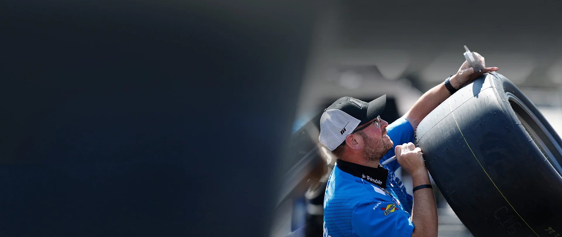 Man in a blue Trimble shirt with Sunoco logo and black cap measures a Goodyear tire tread with a depth gauge, highlighting AECO precision.