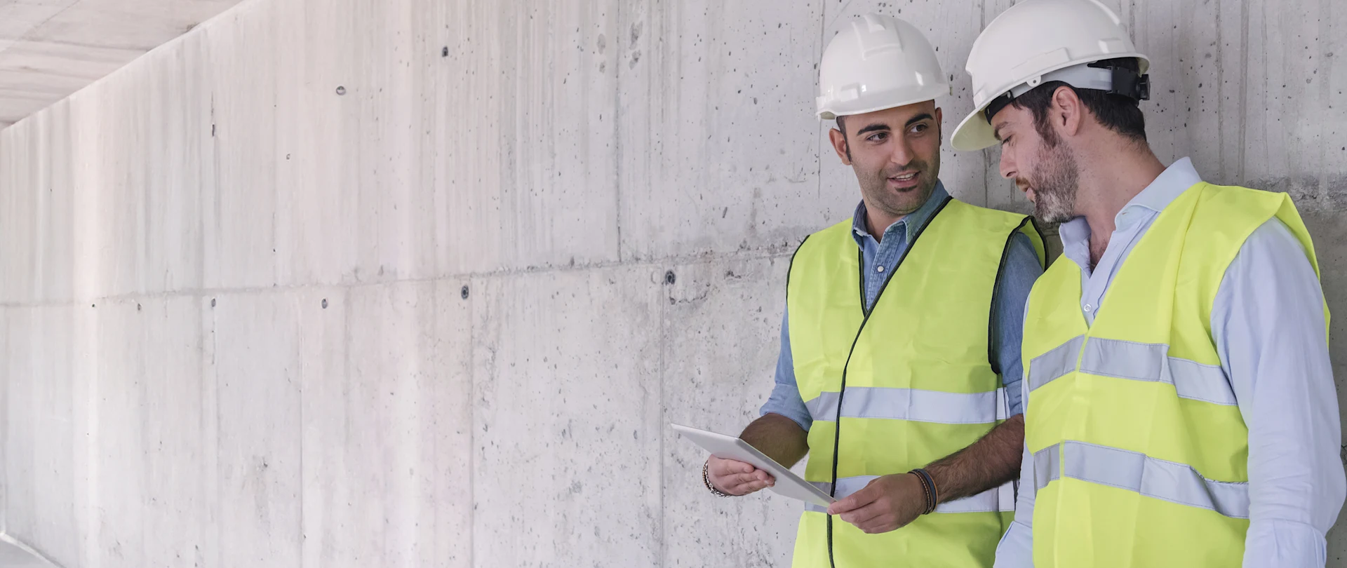 Two construction professionals in hard hats and safety vests look at a tablet while standing in a concrete building.