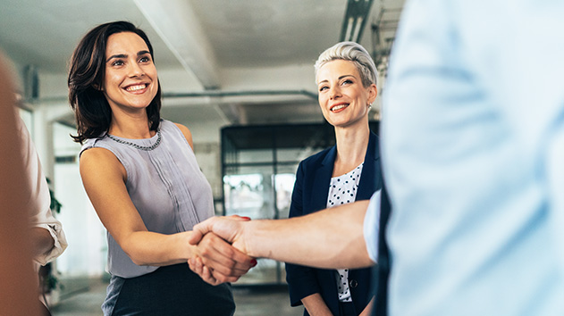 A smiling woman in a sleeveless top shakes hands with a colleague in a bright, modern office as another woman smiles in the background.