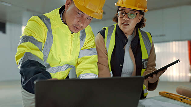 Two construction workers in hard hats and high-visibility vests collaborate over a laptop and tablet at a building site.