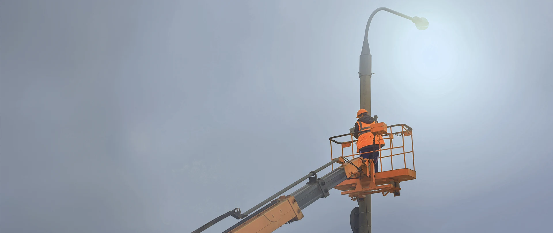 A maintenance worker in a lift bucket repairs a street lamp. A Trimble logo is visible on the yellow hydraulic boom against a hazy sky.