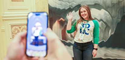 At a Trimble conference, a woman in a 3D EC shirt waves in front of a structural mural, as seen through a smartphone camera.