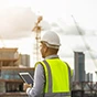 A construction worker in a safety vest and hard hat uses a tablet at a job site with a tower crane and city skyline in the background.