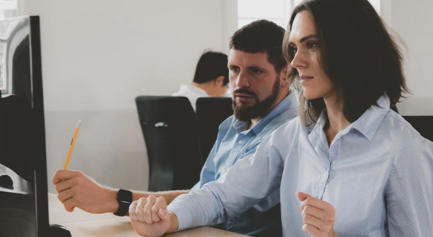 A man with a beard and a woman in blue shirts look at a computer monitor in an office. The man points with a yellow pencil.