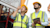 Two construction workers in yellow hard hats and high-visibility safety vests look upward while standing next to heavy machinery.