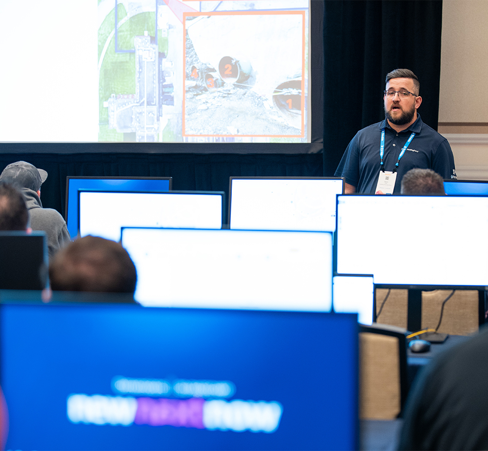 A presenter stands before a classroom of attendees using monitors to follow a technical demonstration at the Trimble Developer Summit.