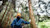 Personne portant une casquette jaune et une veste bleue qui se tient dans une forêt et touche un tronc d'arbre.