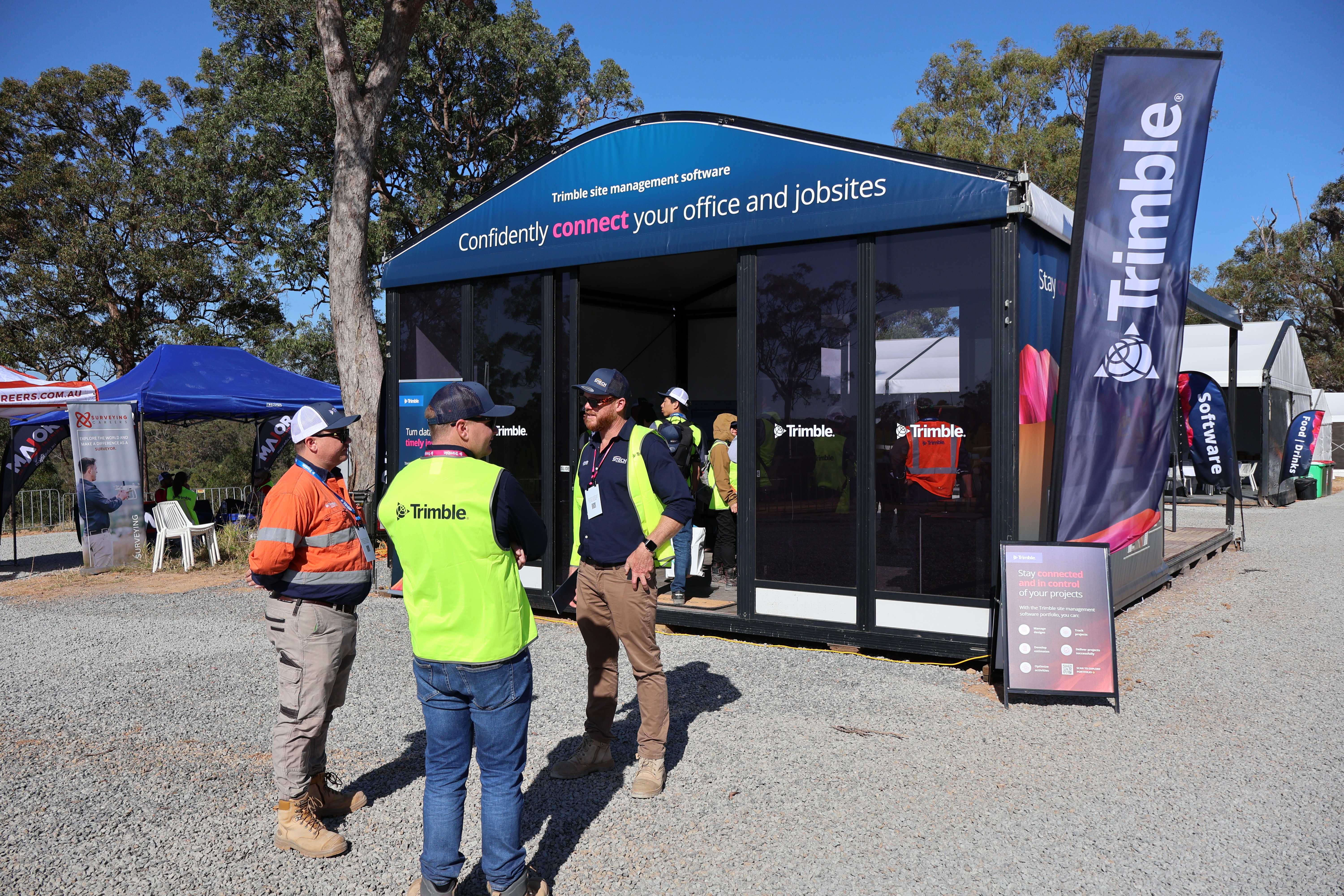 Three men in high-visibility safety gear talk outside a Trimble exhibition booth for site management software at an outdoor industry event.