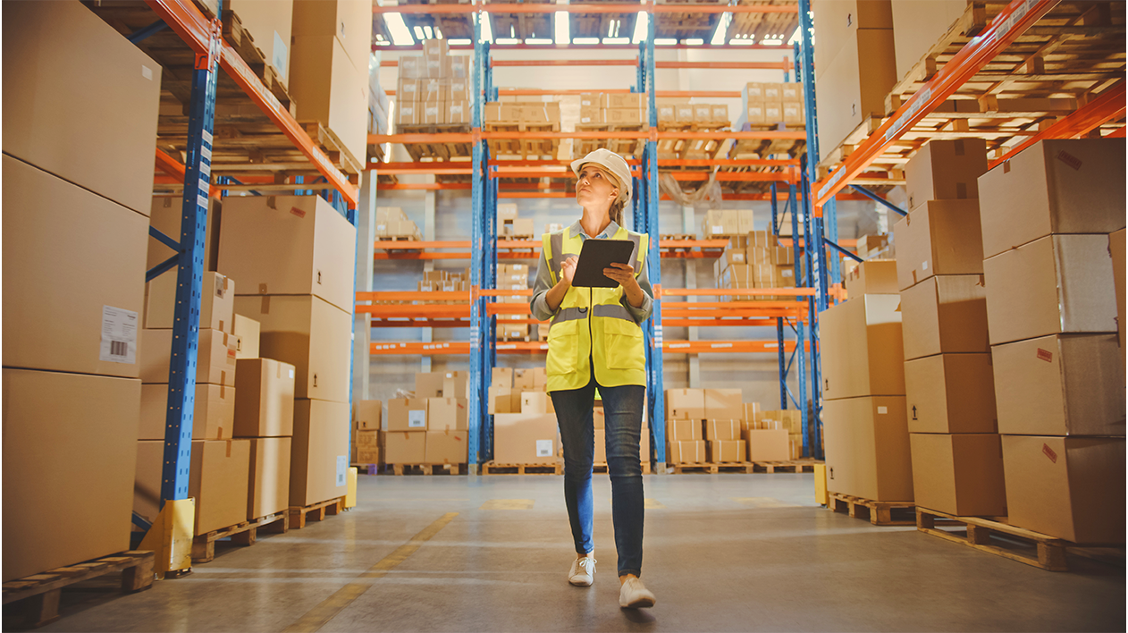 Woman wearing hard hat and yellow safety vest is taking inventory of stacked boxes in warehouse