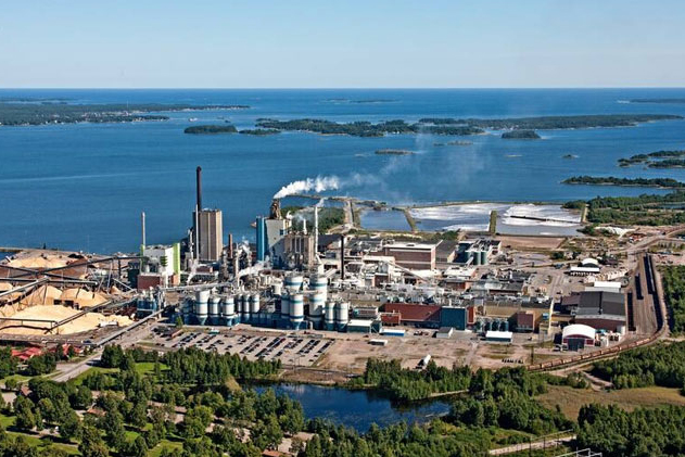 Aerial view of an industrial plant by a large body of water, emitting white smoke. Greenery contrasts with the structures.