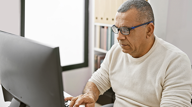 A man wearing glasses and a light-colored sweater looks at a computer monitor in a professional office with a bookshelf in the background.