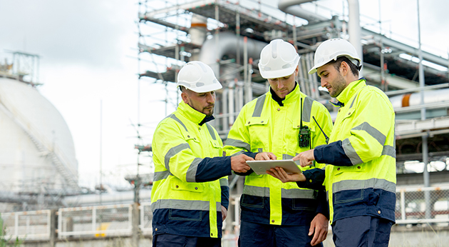 Three industrial workers in high-visibility jackets and hard hats review data on a tablet at a refinery site.