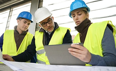 Three construction professionals in hard hats and safety vests review project blueprints and a Trimble tablet at a job site.