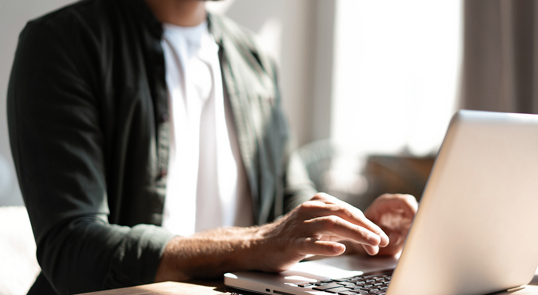 A man in a green button-down shirt typing on a laptop at a wooden desk in a bright, sunlit indoor setting.