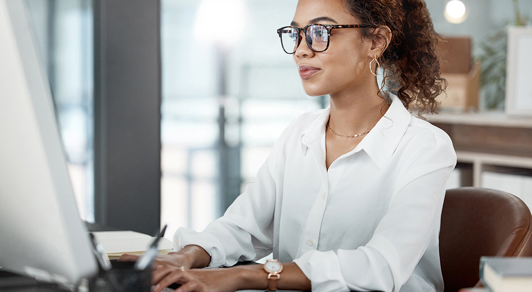 A professional woman with curly hair and glasses works at a computer in a modern office setting.
