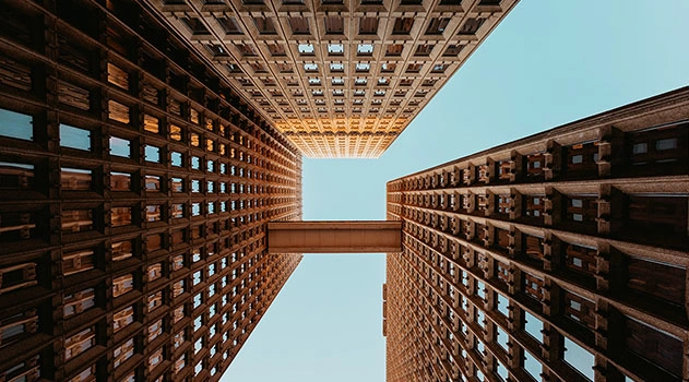 A low-angle view looking up at three tall buildings with grid-patterned facades and a connecting skybridge against a clear sky.
